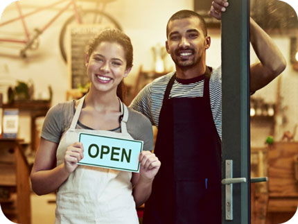 Young couple standing at front door of their small business holding a sign with the word “OPEN”